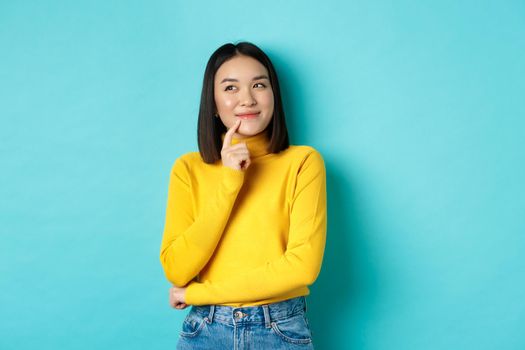 Beautiful korean girl thinking, imaging and smiling, looking at upper left corner and daydreaming, standing against blue background.