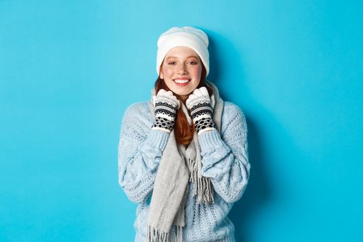 Winter and holidays concept. Cute redhead girl in white beanie and gloves smiling at camera, looking delighted, standing against blue background.
