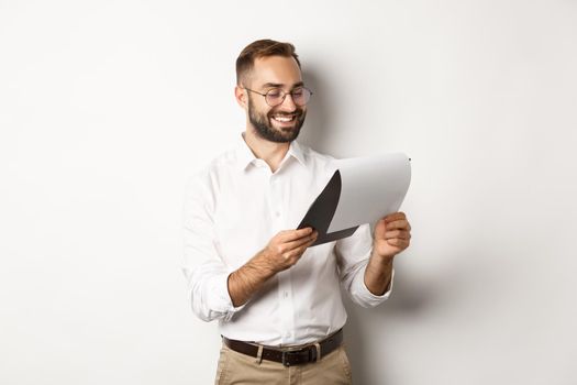 Businessman looking satisfied at documents, reading report and smiling, standing over white background.