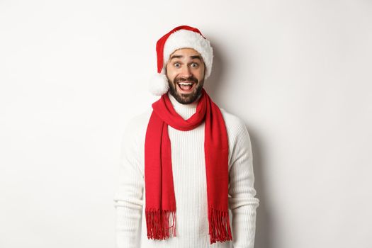 Christmas holidays. Bearded man looking surprised at camera, wearing santa party hat and red scarf, standing against white background.