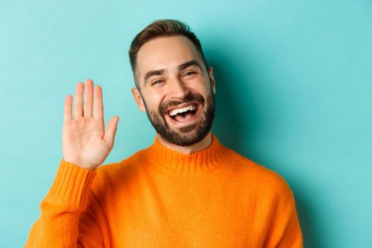 Close-up of friendly young man smiling, waving hand to say hello, greeting you, standing over light blue background.