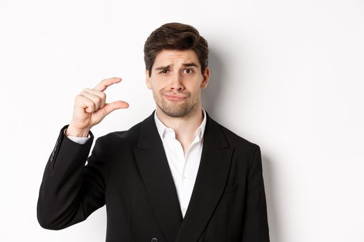 Close-up of handsome businessman in trendy suit, showing something small and looking disappointed, standing against white background.