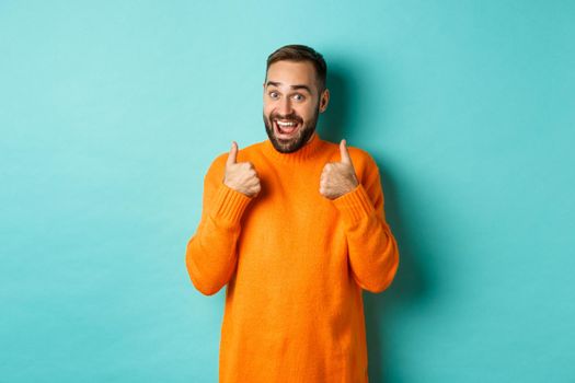Excited young man with beard, showing thumbs up in approval, praise or recommend, standing over light blue background.