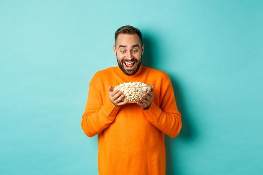 Handsome bearded man looking with exctied and happy at popcorn bowl, ready to watch movie, standing over white background.