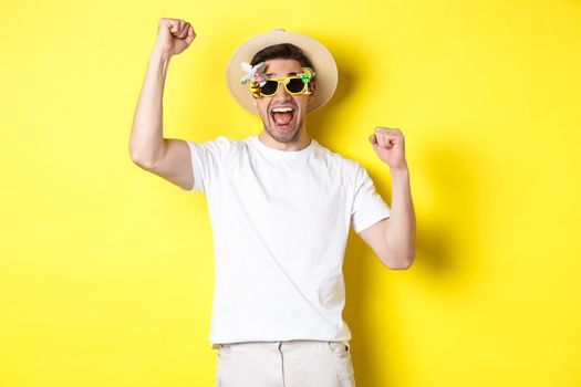 Concept of tourism and lifestyle. Happy man winning trip to resort, shouting yes and raising hands up, triumphing, wearing sunglasses and summer hat, yellow background.