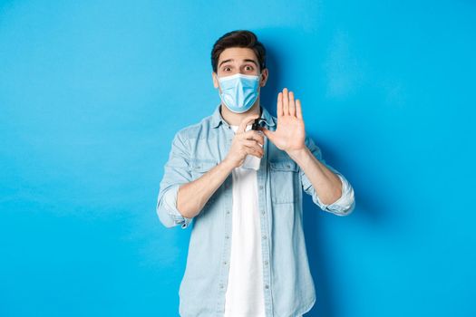 Concept of covid-19, pandemic and social distancing. Cheerful guy in medical mask showing how to disinfect hands with sanitizer, using antiseptic, preventing virus spread, blue background.