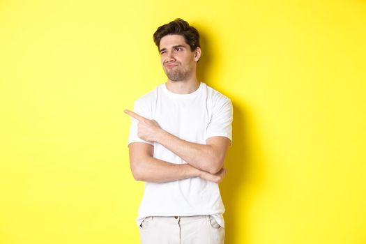 Unimpressed young man complaining, pointing and looking with skeptical face at bad product, standing against yellow background.