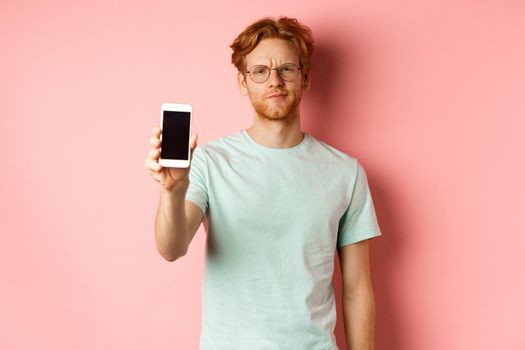 Disappointed male model frowning, showing smartphone screen, standing over pink background.