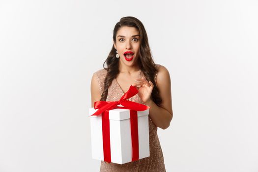 Merry Christmas. Image of attractive woman in luxury dress, open gift and looking surprised, receive new year present, standing over white background.