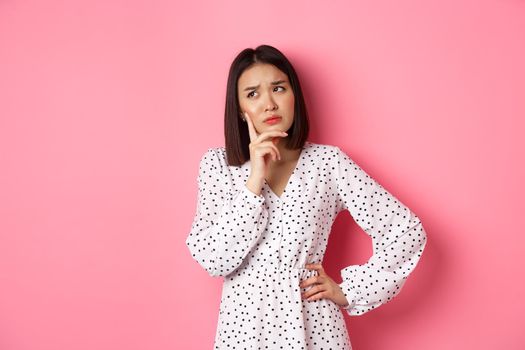 Concerned young asian female model making decision, seriously thinking, looking at upper left corner and choosing, standing over pink background.