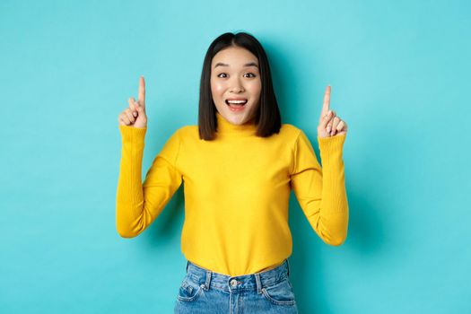 Shopping concept. Impressed young korean woman showing advertisement on top, pointing fingers up and gasping amazed, saying wow, standing over blue background.