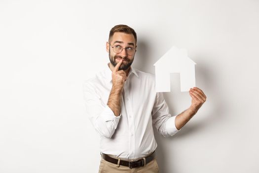 Real estate. Man thinking while searching for apartment, holding paper house model, standing over white background.