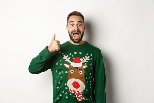 Winter holidays and christmas. Excited caucasian guy showing thumb up and looking amazed, standing in green sweater against white background.