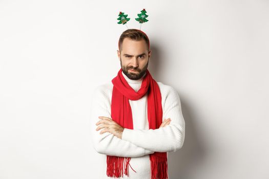 Christmas party and celebration concept. Suspicious young bearded man looking doubtful, wearing funny accessory hat, standing against white background.