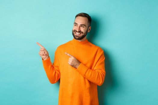 Attractive bearded male model in orange sweater, smiling satisfied and pointing fingers left, standing over turquoise background.