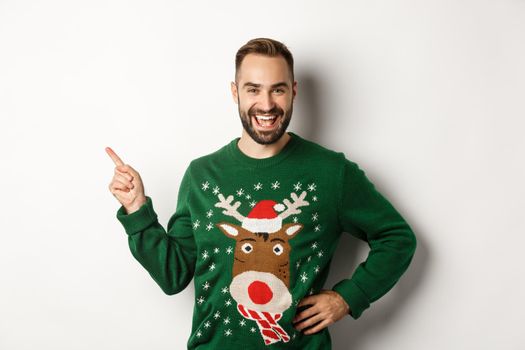 New year celebration and winter holidays concept. Handsome bearded male model in christmas sweater laughing, pointing finger at upper left corner logo, white background.