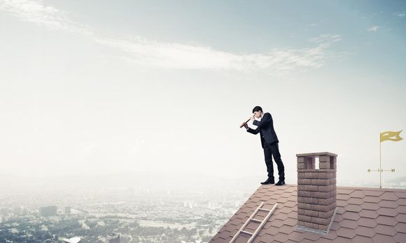 Young determined businessman standing on house roof and looking in spyglass. Mixed media