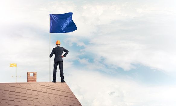 Businessman standing on house roof and holding blue flag. Mixed media