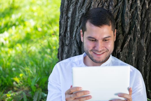 Man using tablet computer under tree in park on sunny day. Handsome guy looking at tablet screen. Happy smiling man sitting on green grass leaning against tree. Digital technology and communication