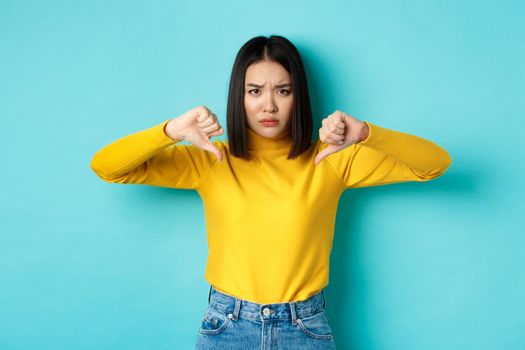 Disappointed asian woman frowning upset, showing thumbs down in dislike and disapproval, standing over blue background, staring at camera.