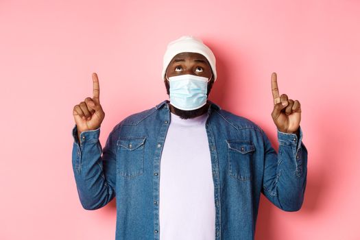 Coronavirus, lifestyle and global pandemic concept. Intrigued Black man seeing advertisement, pointing and staring up at promo offer, standing over pink background.