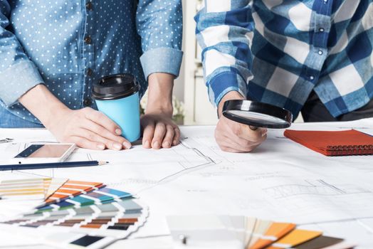 Man carefully studying technical drawing with magnifying glass. Designers working with color swatches and construction blueprint at workplace. Thorough analysis and inspection of design project.