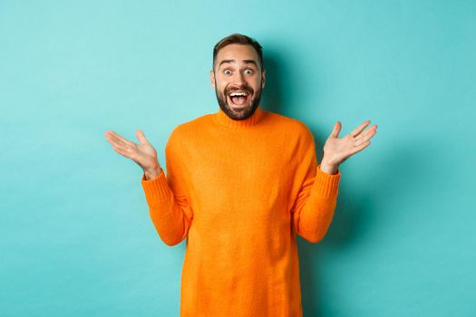 Surprised man raising hands up, looking at something amazing, standing over turquoise background in winter sweater.