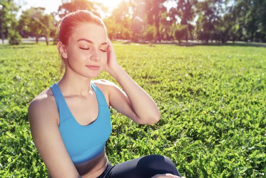 Smiling woman training outdoor at sunny summer day. Morning stretching exercises and healthy lifestyle. Attractive girl doing yoga in park. Young woman with closed eyes sitting on green grass.