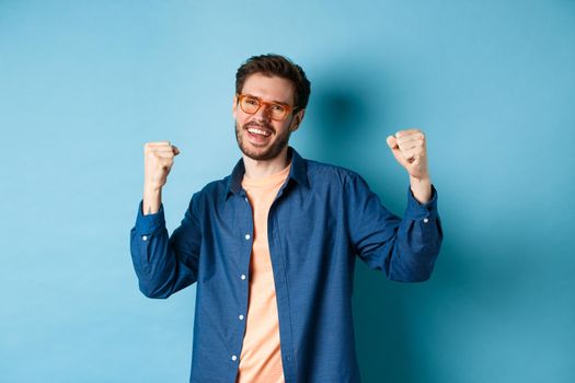 Cheerful young man celebrating, raising hands up and saying yes, wearing eyewear, standing on blue background.