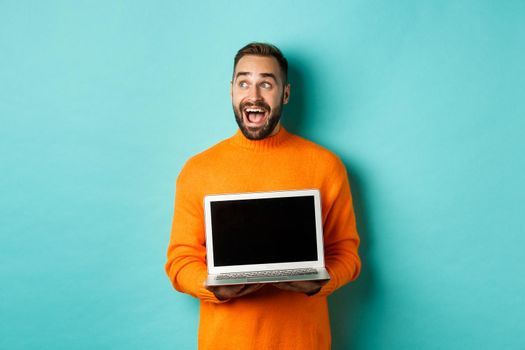 Amazed young man in orange sweater looking at upper left corner, showing laptop screen promo offer, standing over turquoise background.