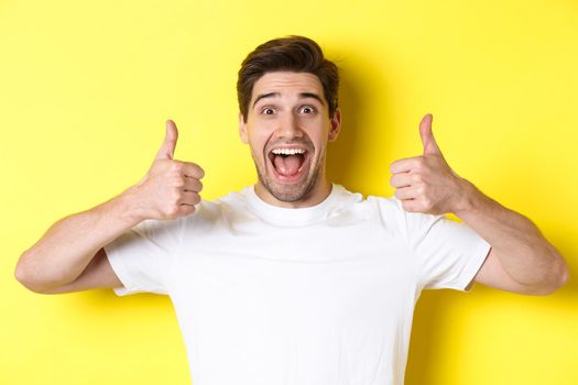 Close-up of handsome young man showing thumbs up, approve and agree, smiling satisfied, standing over yellow background.