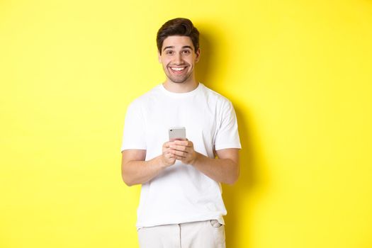 Man smiling and looking happy after reading promo offer on smartphone, standing against yellow background in white t-shirt.