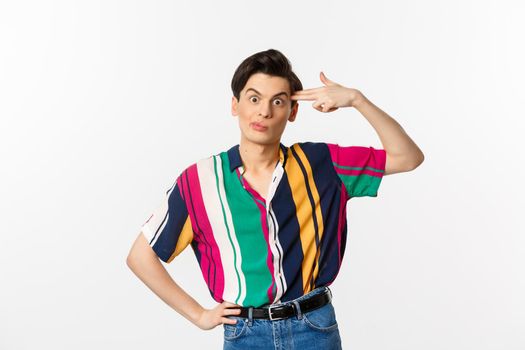 Annoyed gay man making finger gun sign, shooting hismelf from irritation and boredom, standing over white background.