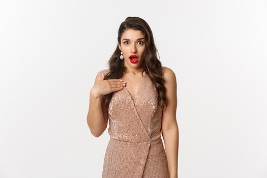 Winter holidays and party concept. Image of attractive woman with red lips, wearing glamour dress and pointing at herself with shock, looking surprised, standing over white background.