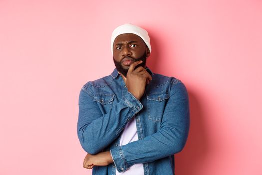 Doubtful african american man staring at camera with disbelief, standing skeptical and thinking, pink background.