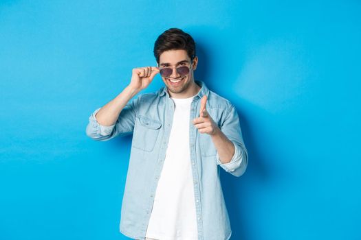 Handsome cheeky guy in sunglasses, smiling and pointing finger gun at camera, flirting with you, standing over blue background.