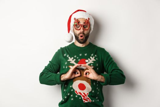 Christmas holidays, celebration concept. Happy guy in Santa hat and party glasses making fun of funny sweater, standing over white background.