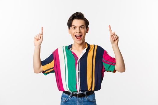 Happy attractive gay man pointing fingers up, smiling excited, showing top offer, standing over white background.