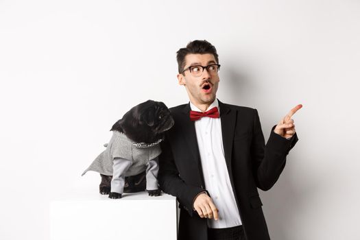 Animals, party and celebration concept. Amazed young man and black dog in costumes staring right at copy space, standing against white background.