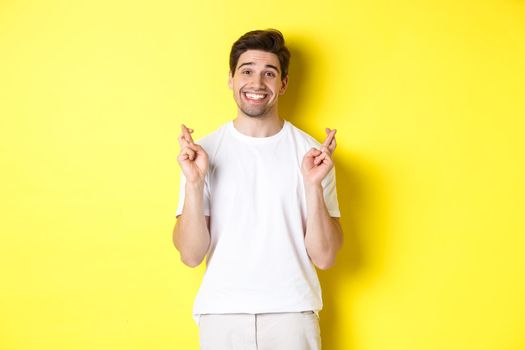 Hopeful smiling man holding fingers crossed, making wish or praying, standing over yellow background.