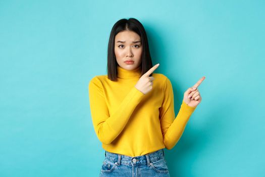 Shopping concept. Disappointed korean girl looking gloomy, asking to buy this, pointing fingers at upper right corner and staring sad at camera, blue background.