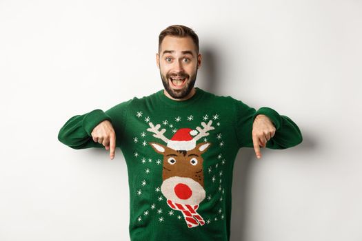 Winter holidays and christmas. Excited bearded man in green sweater, pointing fingers down and looking amazed, standing over white background. Copy space