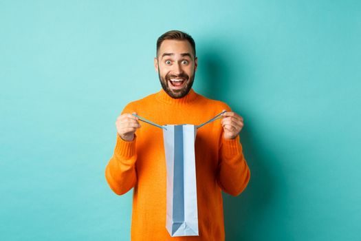 Surprised man open shopping bag and looking amazed, receiving gift on holiday, standing over turquoise background.