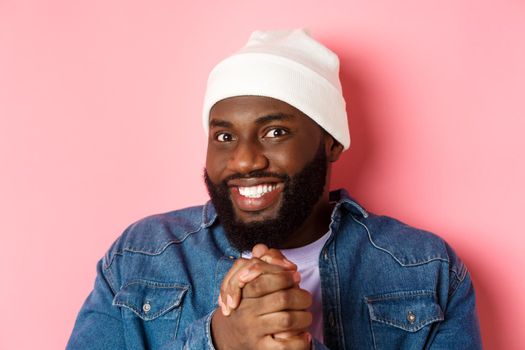 Close-up of devious and happy african-american man smiling satisfied, clasp hands think something good, expect profit, standing over pink background.