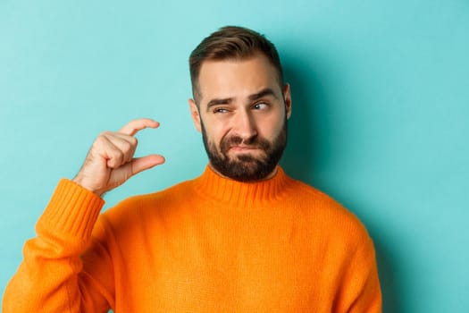 Close-up of caucasian man showing small or little with fingers, standing disappointed over light blue background.