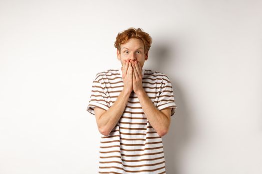 Surprised and excited man with curly red hair checking out special promo offer,staring fascinated and amazed, covering mouth with hands, standing over white background.