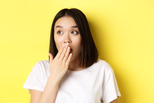 Close up of pretty asian girl looking shocked, gasping and covering mouth with hand, stare left at promotion, standing over yellow background.