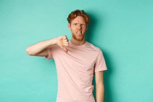 Disappointed redhead man showing thumbs-down and tongue, epxress dislike and disapproval, standing over turquoise background.