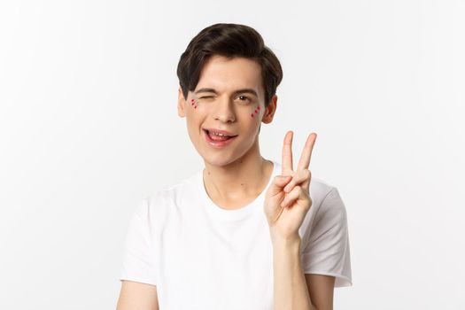 People, lgbtq community and lifestyle concept. Happy and cute gay man with glitter on face, showing peace sign and smiling, celebrating pride holiday, white background.