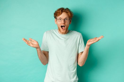 Surprised handsome man with red hair, wearing glasses, spread hands sideways and gasping in awe, looking wondered at camera, standing over turquoise background.